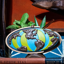 Ceramic tiki mug shaped like a globe in green and blue with brown accents, surrounded by mint and pineapple leaves.