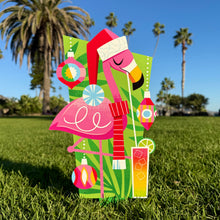 Colorful flamingo yard stake with holiday decorations, vibrant design, standing on grassy lawn against blue sky.